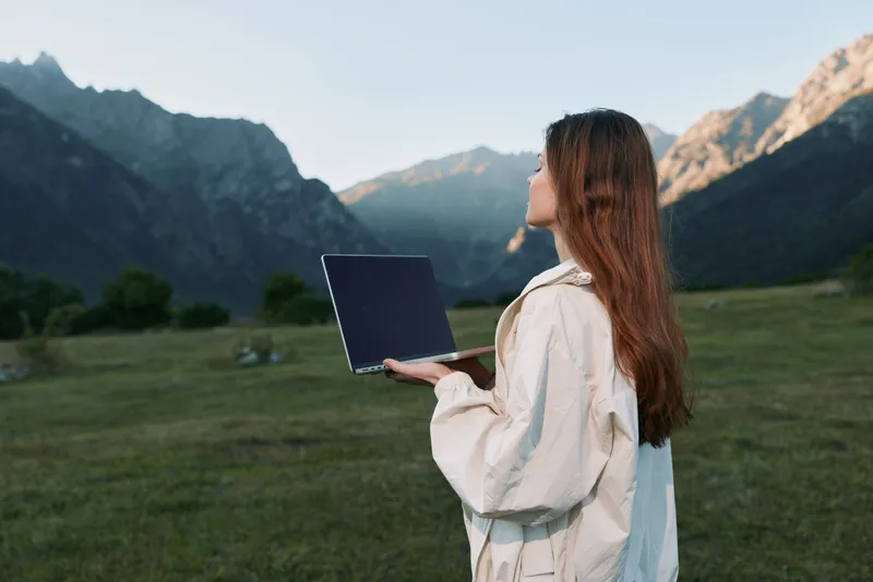 A focused woman outdoors holds a laptop in a scenic valley with distant mountains, conveying remote work, concentration, and a serene, productive moment in nature.