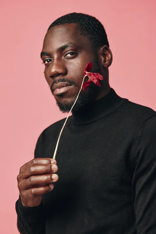 African man with dark skin holding red flower near face in creative conceptual visual style. Portrait with black turtleneck and pink background for artistic expression and beauty.