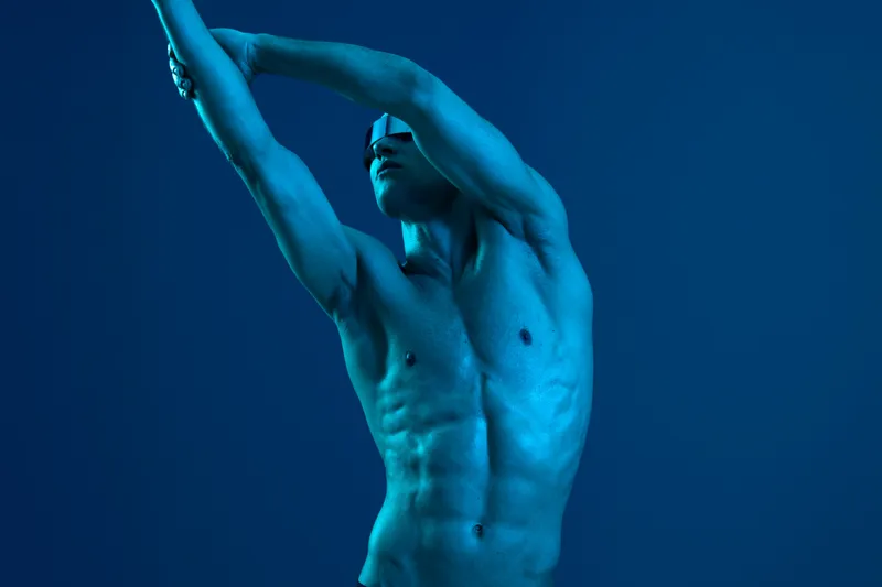 Athletic man posing dramatically in blue light, showcasing strength and expression, emphasizing the beauty of movement. A striking black and white image.