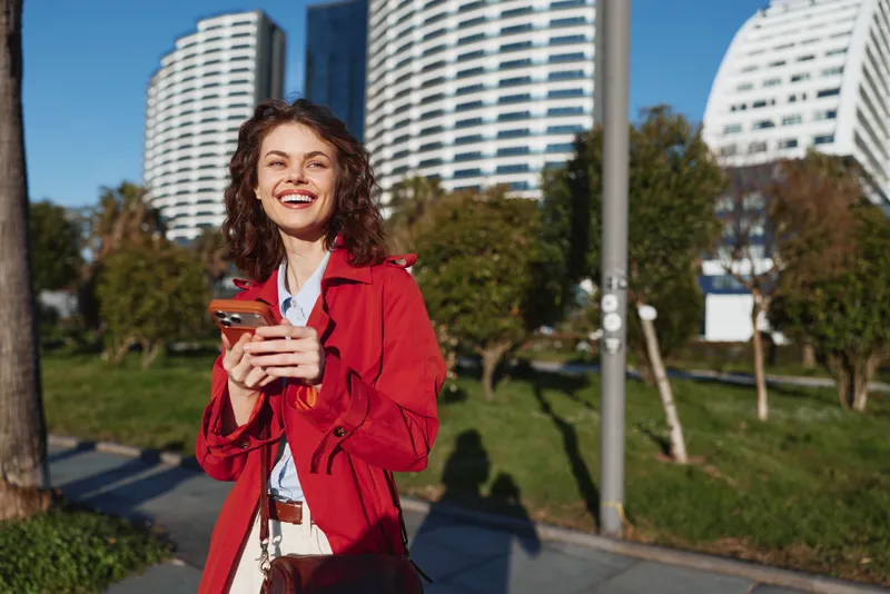 Joyful young woman in a red jacket outdoors, smiling while holding a smartphone, urban park setting with modern buildings in the background and soft shadows