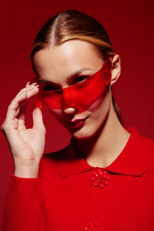 Stylish woman in red sweater and red translucent sunglasses poses against red background. Fashion portrait with bold color contrast and confident expression. Concept of modern style and elegance.