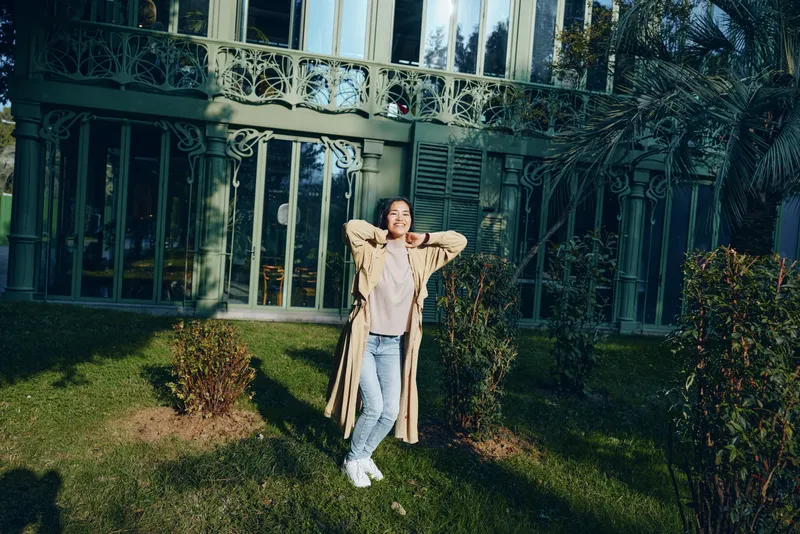 Woman standing in front of a glass building with a lush garden, relaxed pose, beige coat and jeans, casual street style against modern architecture and bright sunshine