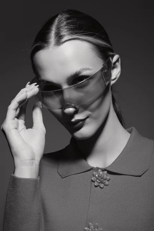 Young white woman with fair skin wears sunglasses and a vintage sweater, posing with confident expression in studio lighting. Fashion and style concept.