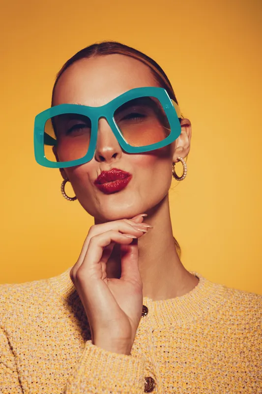 Young woman with oversized turquoise glasses and red lipstick poses with hand near chin against yellow background. Fashion portrait with playful expression and bright studio lighting.