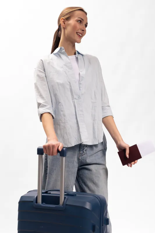 Young woman in casual clothes holding suitcase and passport with ticket, ready for travel. Smiling traveler with ponytail on white background, concept of vacation and trip preparation.