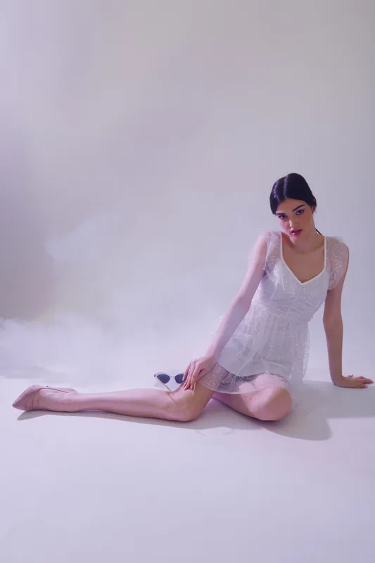 Young woman in a white sheer dress sits on the floor with legs extended. Soft lighting highlights her beauty and fashion style in a minimal studio setting.
