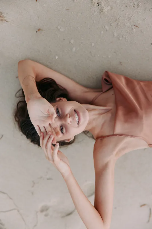 A relaxed portrait of a woman lying on warm sand, wearing a peach dress, with a soft smile and gentle lighting that highlights skin and fabric texture.