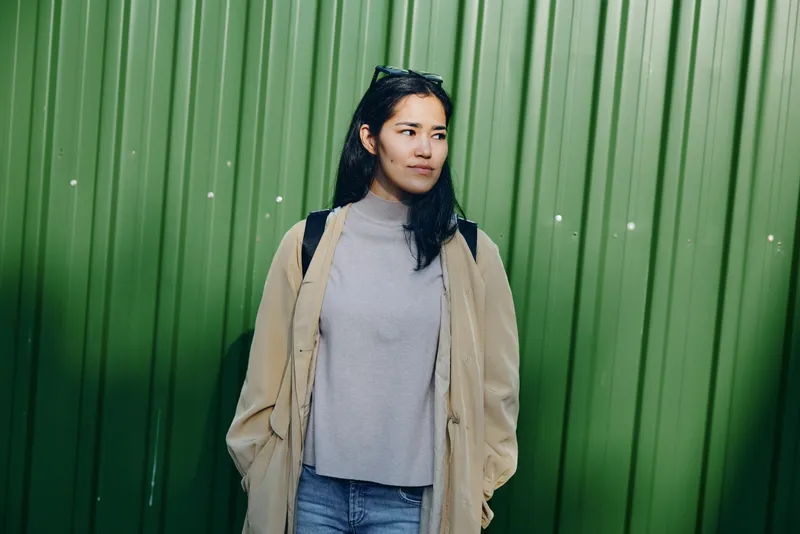 Woman in a beige coat and gray knit top stands outdoors against a vivid green wall, showcasing casual street fashion and contemplative poise in an urban setting.