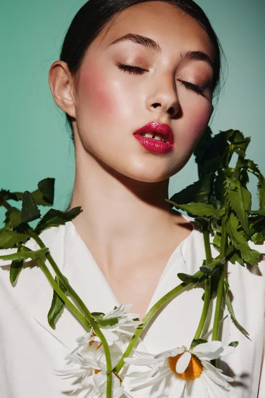 Young woman with glossy red lips and shimmery eyeshadow poses with white daisies against sage green background for beauty and fashion creative concept.