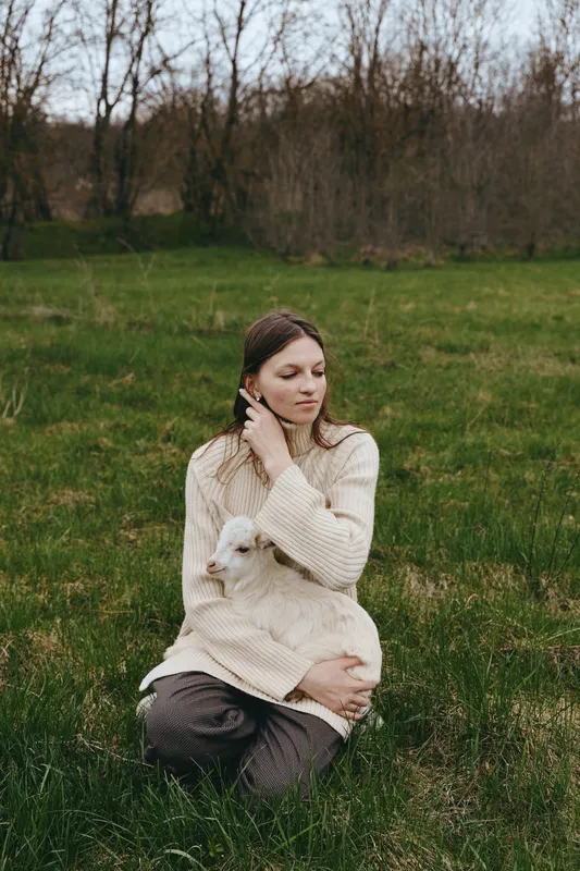 Lifestyle portrait of a young woman sitting in a green field, comforting a small goat kid, wearing a cozy sweater, a calm moment of bond with nature, soft light, rural mood, peaceful and tender.
