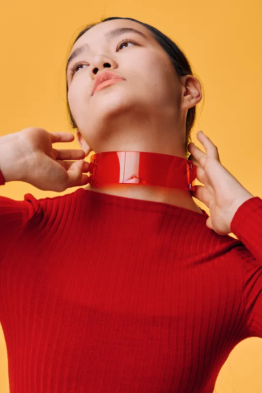 Woman wearing red sweater and shiny red collar looks upwards with hands near neck on yellow background. Portrait of young female with smooth skin and minimal makeup in studio light.
