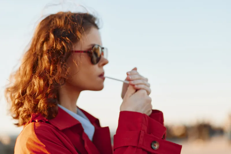 Young woman with curly red hair in a red jacket lights a cigarette, absorbed in the moment with a confident, rebellious vibe against a blurred outdoor backdrop.