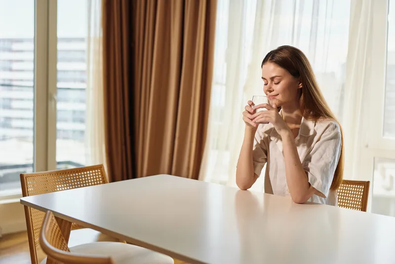 woman with cup at table in morning sunlight smiling with closed eyes, relaxed at home enjoying coffee mug by window and curtains, cozy interior portrait of calm domestic leisure