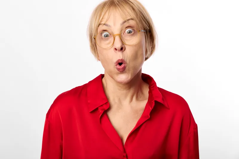 Surprised elderly woman in red shirt, closeup shock expression, studio portrait with wide open eyes and mouth, plain white background, clean studio backdrop