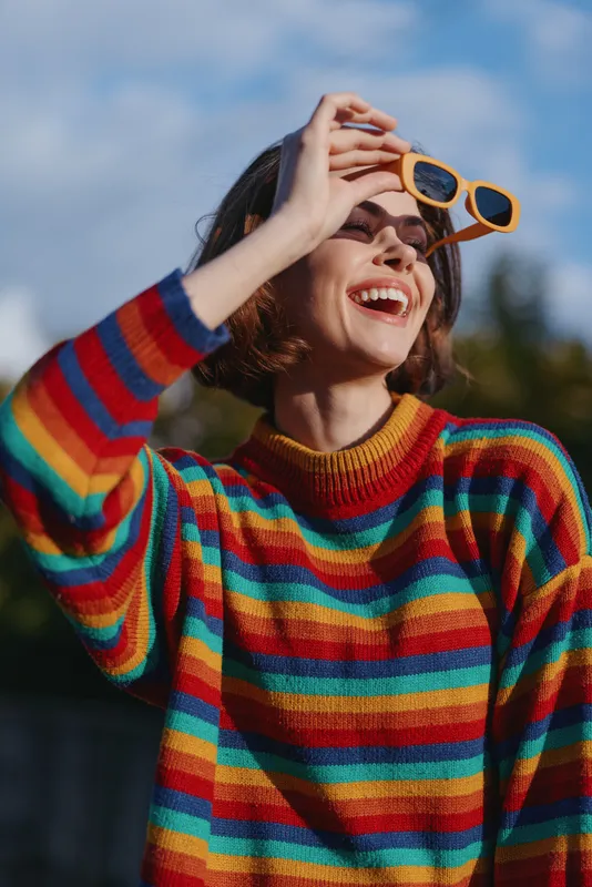 Woman in a colorful striped sweater smiling outdoor with short hair, holding sunglasses and laughing — casual outfit lifestyle portrait with joyful travel and fashion vibe.