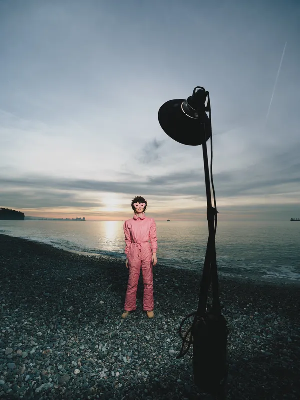 Pink jumpsuit wearer stands on a pebbled beach at sunset with a large studio light nearby, creating dramatic lighting for a fashion or artistic portrait by the water
