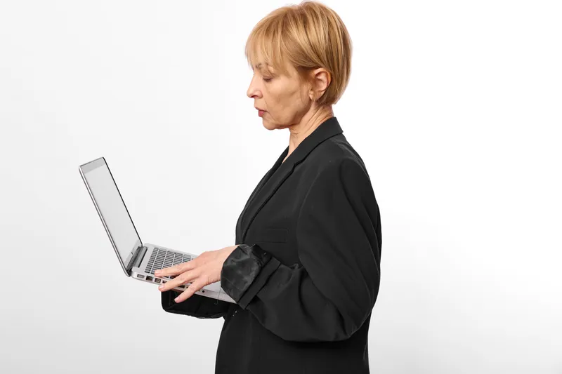 Senior woman with laptop in a clean studio background, typing on the keyboard, focused daily remote work concept, office communication and technology