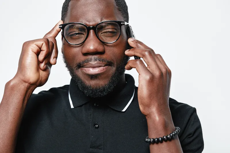 African American man with dark skin wearing glasses and black polo shirt talking on smartphone with thoughtful expression isolated on white background.