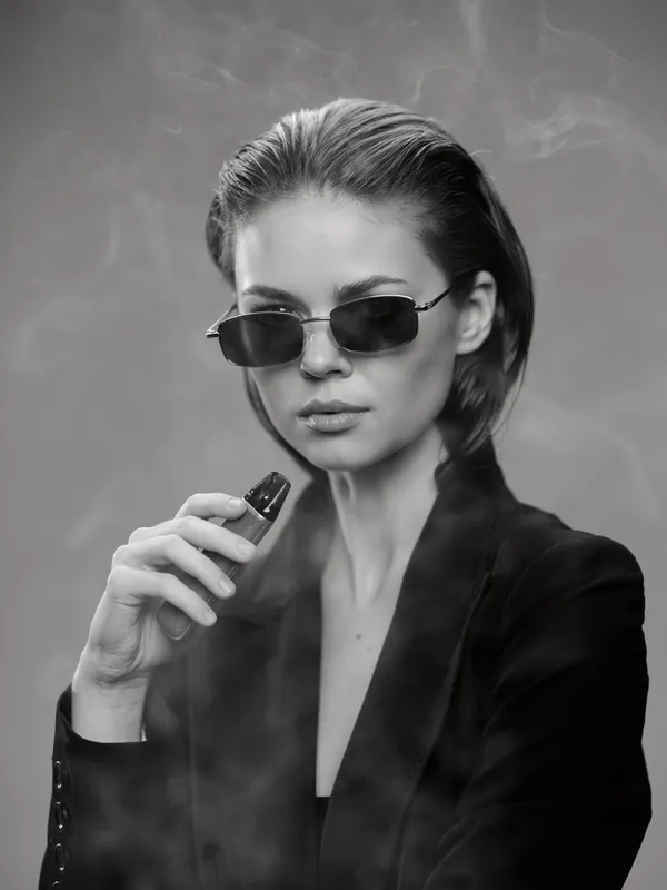 Black and white portrait of stylish woman vaping with sunglasses, confident pose, and smoky atmosphere