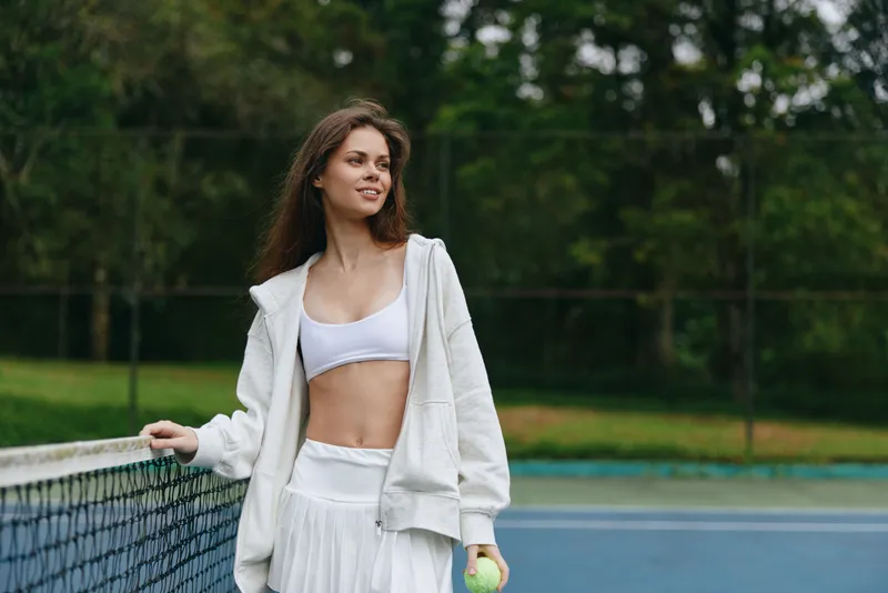 Young woman in sporty clothing smiling while holding a tennis ball, representing an active lifestyle and healthy sports activities in a lush green tennis court