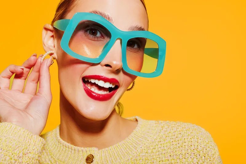 Smiling woman with bright red lipstick wears large turquoise sunglasses and yellow sweater against vibrant yellow background. Concept of fashion, summer style, and joyful mood.