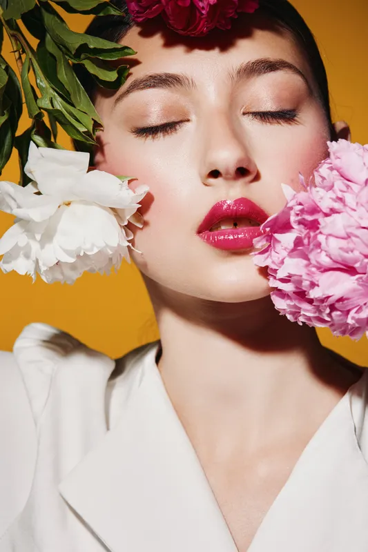 Close-up portrait of a young woman with bright topical makeup and closed eyes framed by vivid colorful flowers on a yellow background. Concept of beauty fashion creativity.