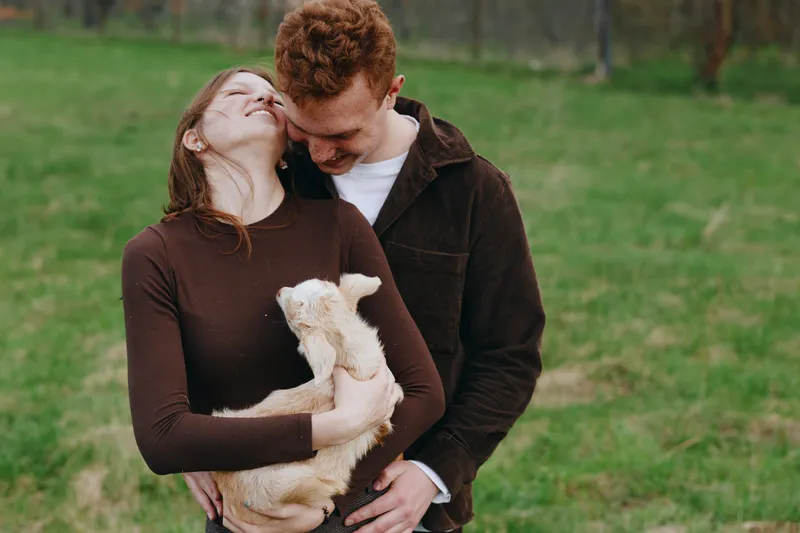 Joyful couple outdoors in a green field cuddle a goat kid, sharing affection and smiles, a cozy moment of bonding with the playful animal companion in a serene rural setting.