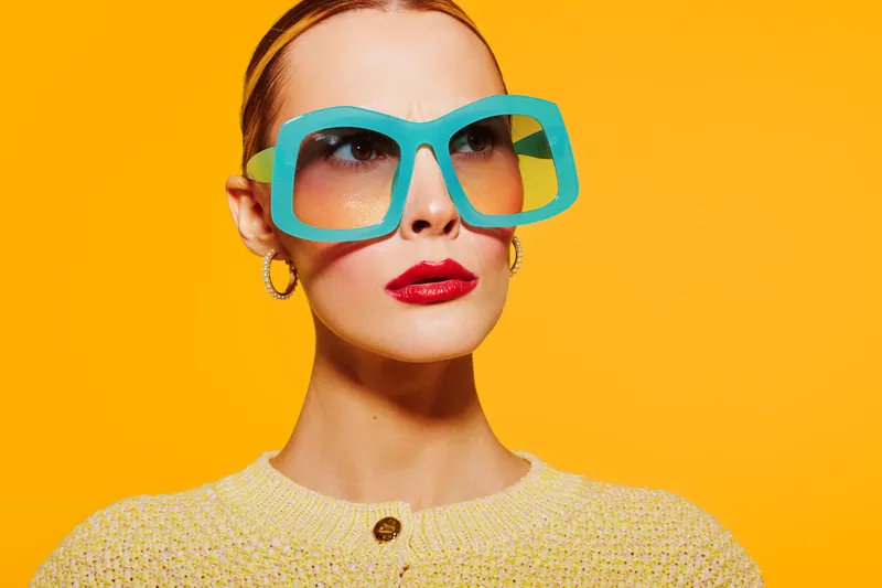 Stylish woman with large blue sunglasses and red lipstick poses against bright orange background. Fashion portrait with vibrant colors and confident expression. Concept of modern style and beauty.