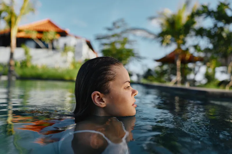Young woman with wet hair relaxing in outdoor pool surrounded by tropical plants and wooden structures. Concept of lifestyle, leisure, and summer vacation in natural light.