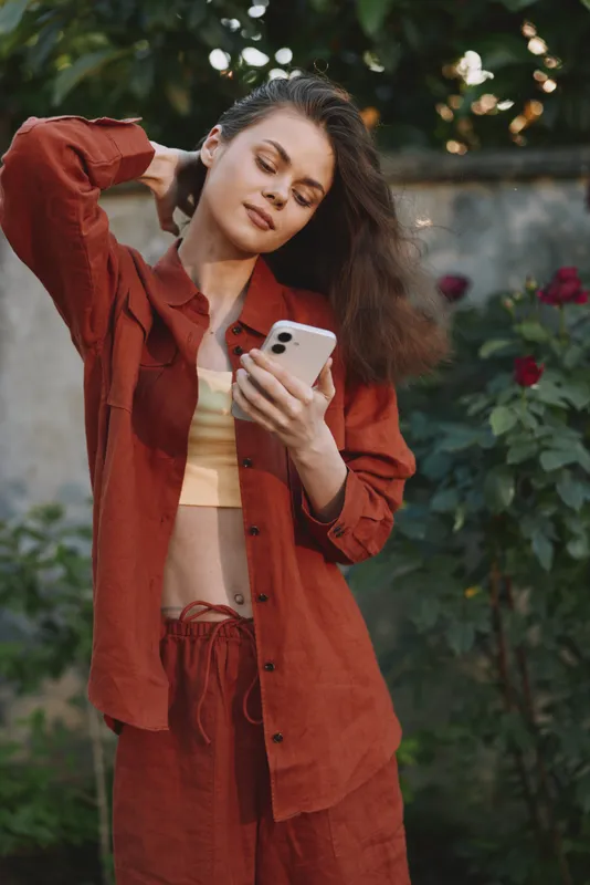 A stylish woman stands outdoors in a rust red outfit, looking at her phone with a calm smile, capturing everyday selfcare and casual fashion moments