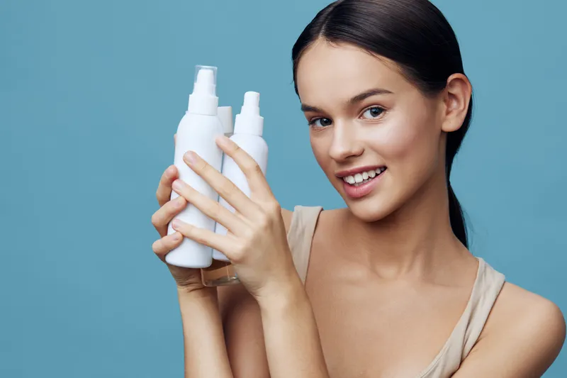 Young woman holding skincare products, showcasing healthy skin and confidence, against a soft blue background, emphasizing beauty and self care
