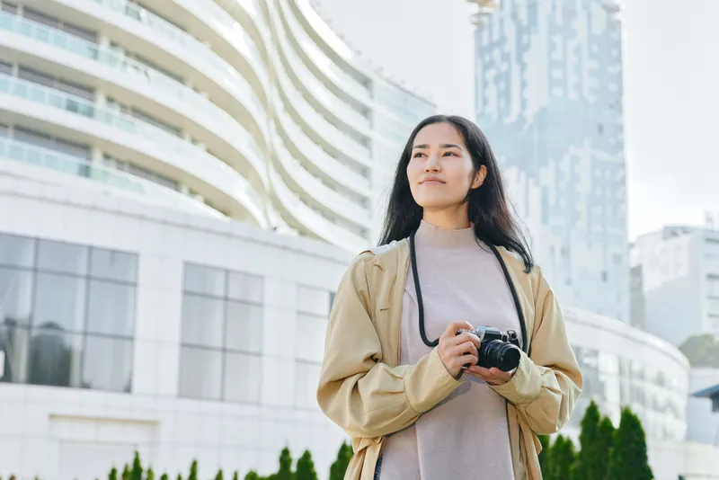 Woman with a camera stands outdoors in a modern cityscape, exploring architecture and street life, casual jacket, urban vibe, travel focus and photography, candid and serene expression
