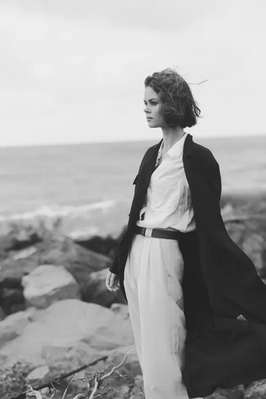 Fashion, woman, wind, coastal, elegance, coat. A monochrome scene shows a stylish figure by the sea in a flowing outerwear over a tailored white suit, conveying confidence, poise and modern