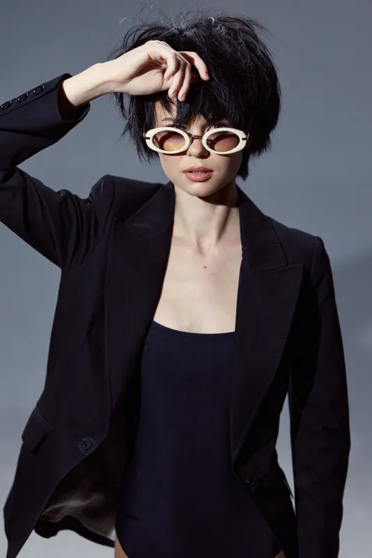 Stylish woman wearing black blazer and sunglasses with short hair, posing confidently against gray background.