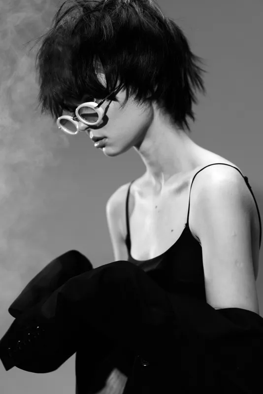 Black and white portrait of a stylish woman with short hair wearing round sunglasses and elegant dark clothing, expressing a thoughtful mood.