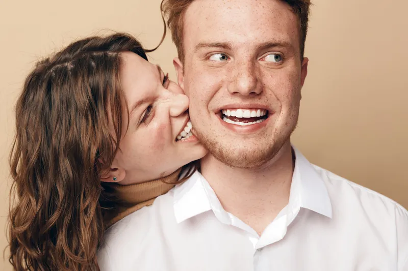 Happy young couple in casual white shirts share joyful moment, woman playfully bites man's cheek. Close-up on beige background. Concept of love, affection, and relationship happiness.