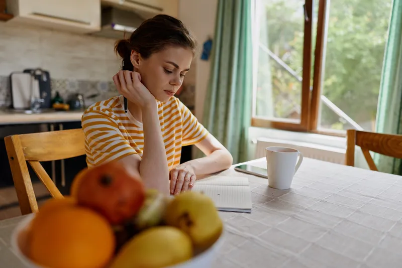 Woman sits at a bright kitchen table, lost in thought, with a fruit bowl and notebook nearby, striped shirt, morning light, domestic setting, a quiet reflective moment.