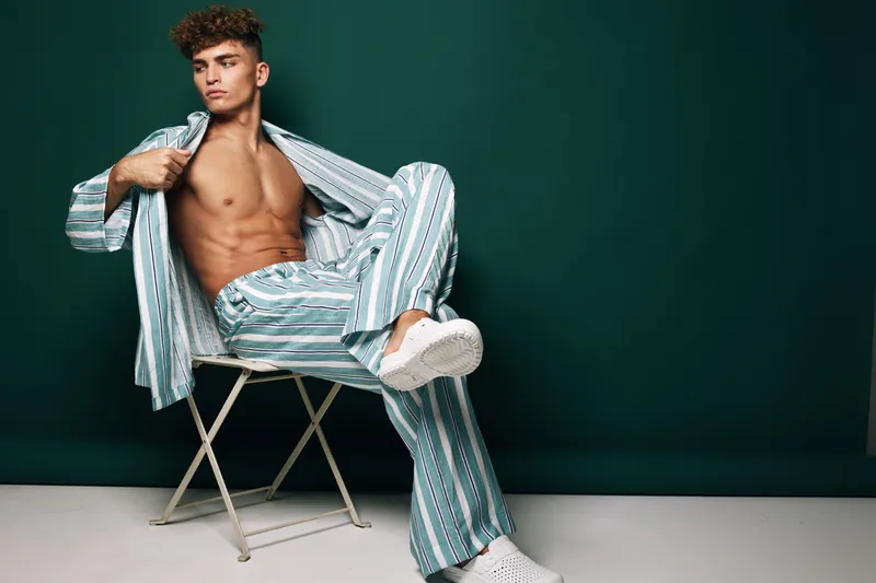 Young man with curly hair sitting on folding chair wearing striped pajamas and white sneakers against dark background. Casual pose with open shirt showing toned body.