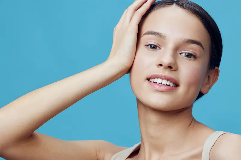 Young woman with a radiant smile and natural beauty on a blue background, embodying confidence and health in a light, casual outfit, promoting self care and positivity