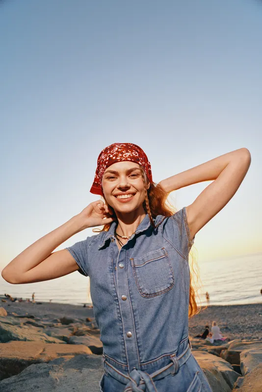 smiling woman in denim dress wearing red scarf enjoys sunny beach sunset with relaxed joyful mood and natural light atmosphere