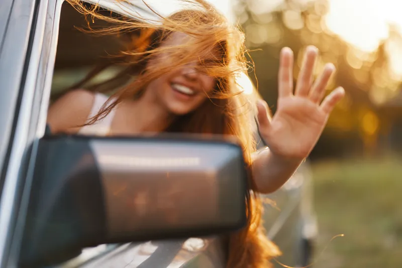 Happy young woman with long hair enjoying a summer road trip, waving from car window with sunlight background, conveying freedom and joy.