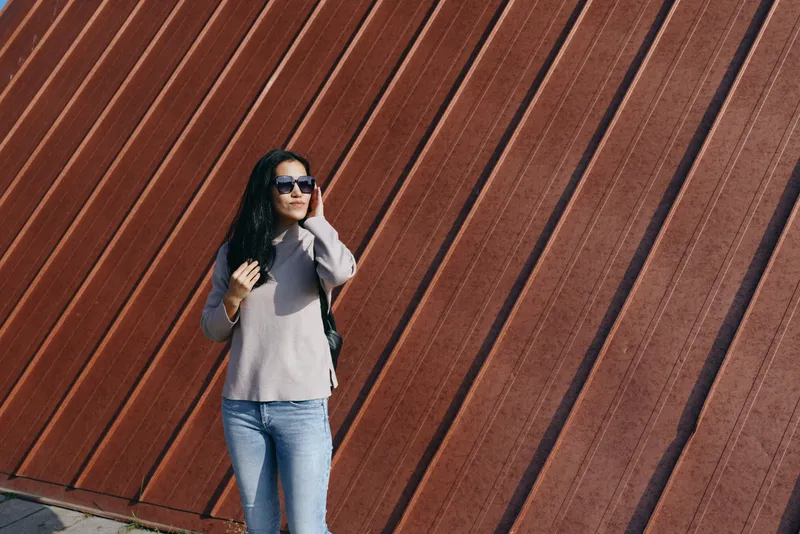 woman fashion outdoors sunglasses backpack casual street urban denim sweater beside a diagonal wall, a confident pose under daylight, showcasing travel style, modern chic and streetwear vibe