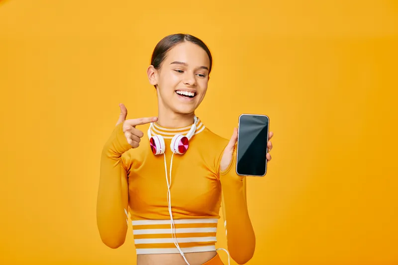 Woman in an athletic orange top with headphones, smiling and showing a smartphone, capturing a fitness lifestyle for sport, training and digital motivation in a bright studio.