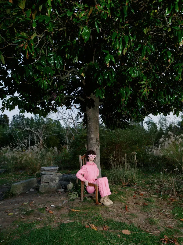 Woman in a pink dress sits on a wooden chair beneath a large leafy tree in a quiet park, enjoying shade and nature in a serene outdoor scene.