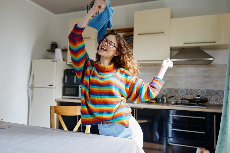 Young woman with curly hair dances playfully in a bright kitchen while hanging laundry, conveying a cheerful, carefree mood and casual home lifestyle.