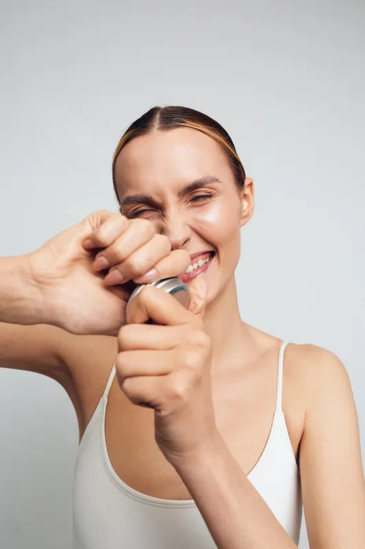 Young woman in white tank top opens a bottle with effort and smiles with closed eyes isolated on light background. Casual lifestyle and positive emotions concept.