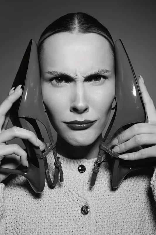 A young caucasian woman with light skin holds two high heel shoes by her face, making a confused expression in black and white portrait.