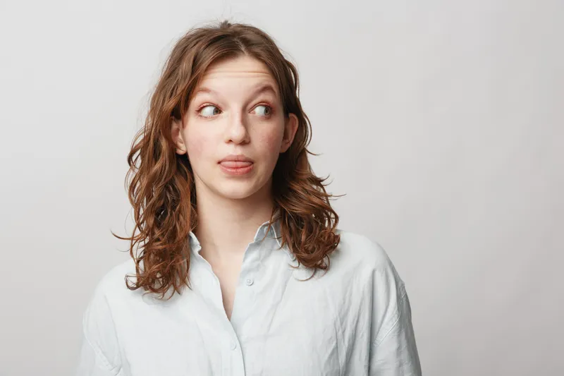 Young woman with curly hair wearing white shirt looks sideways with playful expression sticking out tongue. Studio portrait on light background. Concept of fun, youth, and casual style.