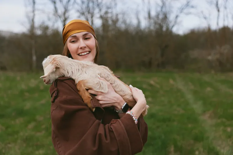 Smiling woman in a brown jacket holds a goat kid in a grassy field, outdoor lifestyle moment with nature, capturing affection, warmth, and gentle care, rustic setting, soft daylight, peaceful mood