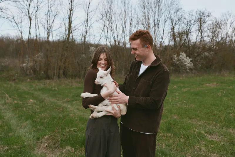 couple lifestyle goat kid A joyful couple outdoors with a playful goat kid, holding the young animal between them in a green field with budding trees, conveying warm affection and wholesome outdoor